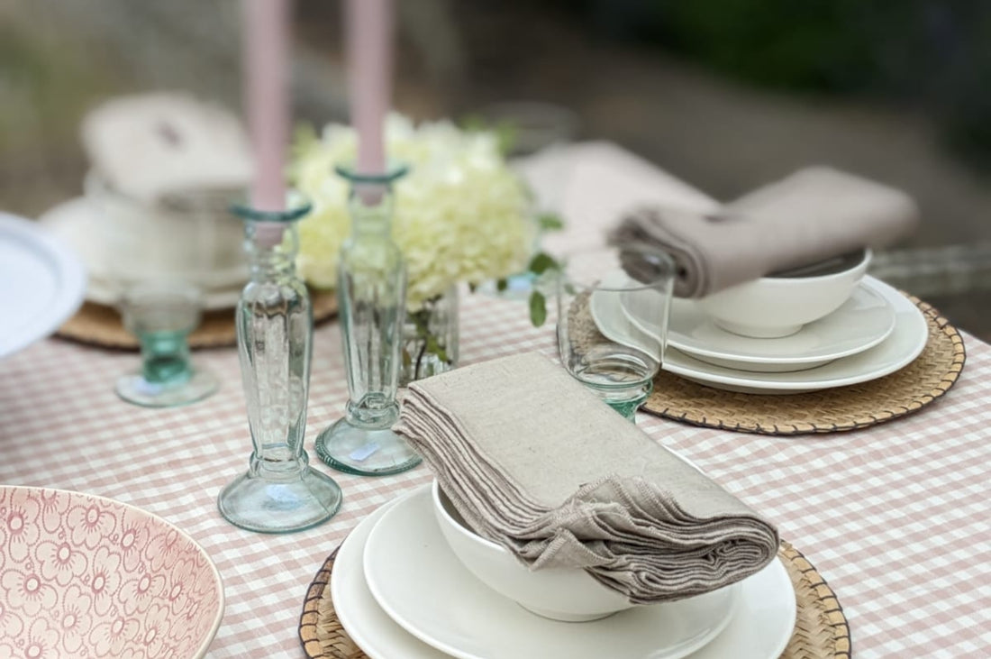 Chalky pink and dusty blue gingham textiles in the form of tablecloths, table runners and seat pads, styled at a garden table, placed alongside flowers and candles.