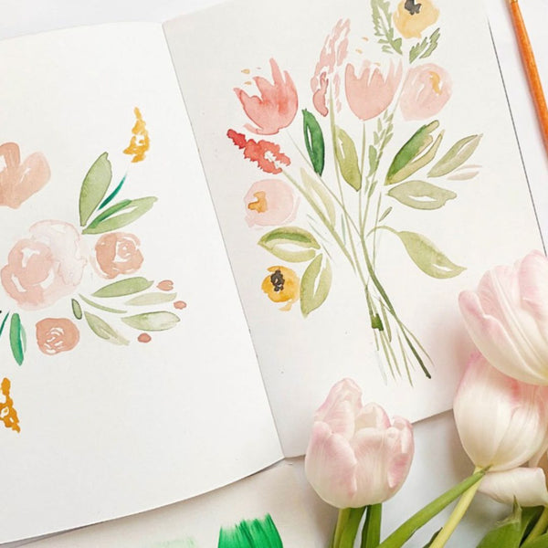 A watercolour painting, within an art book, alongside spring tulips on a table